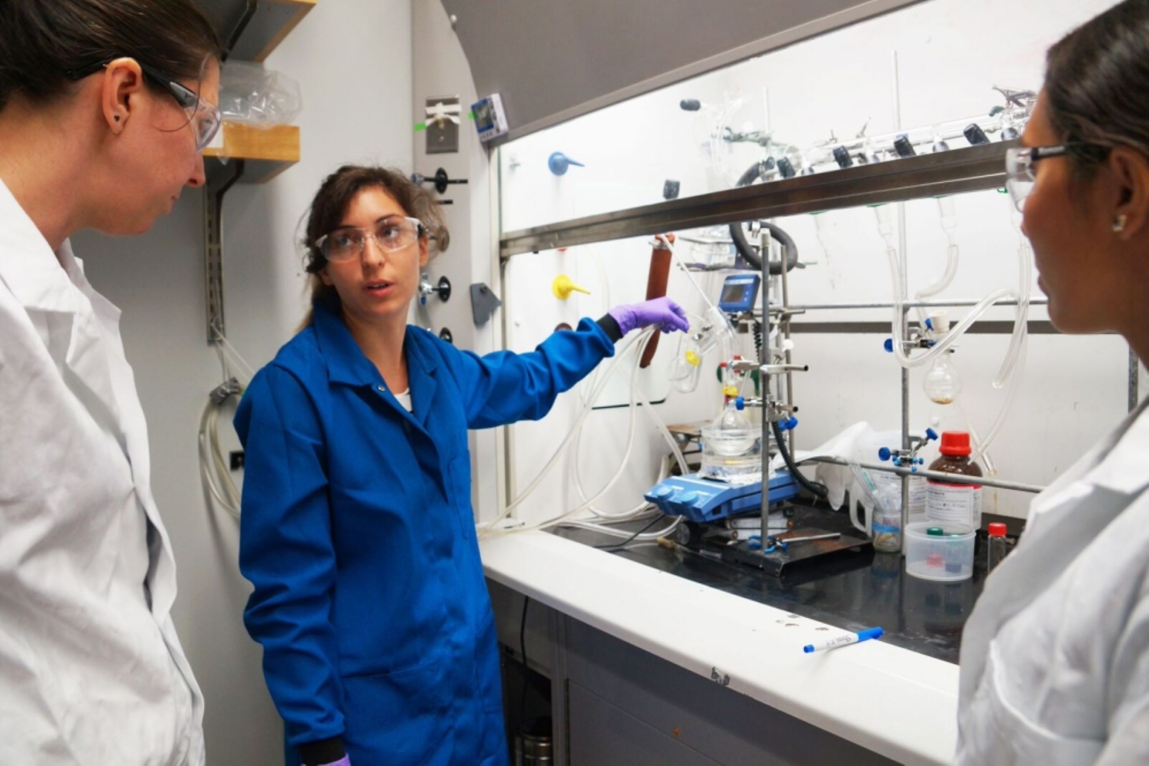 woman wearing blue lab coat, goggles, and gloves showing lab equipment to 2 other women wearing white lab coats and goggles, 